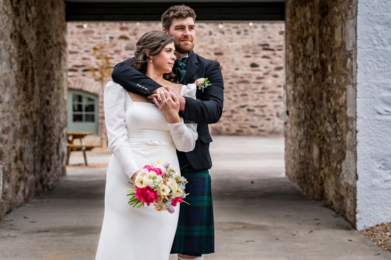 Groom hugs bride from behind while she holds small colourful bouquet in courtyard at Kinclune Steading